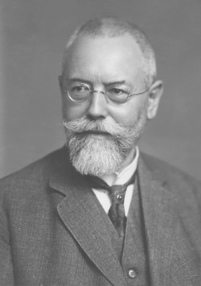 Black-and-white portrait photo. An older man with a white beard and round metal-rimmed glasses, in a suit, looking straight ahead