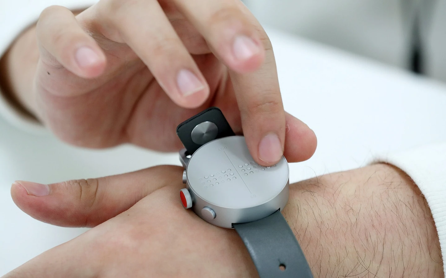 A hand touching a round smart braille watch worn on a wrist with the fingers of the other hand. Braille pins are raised on the watch face. 2017, at Dot's office in Geumcheon-gu, Seoul
