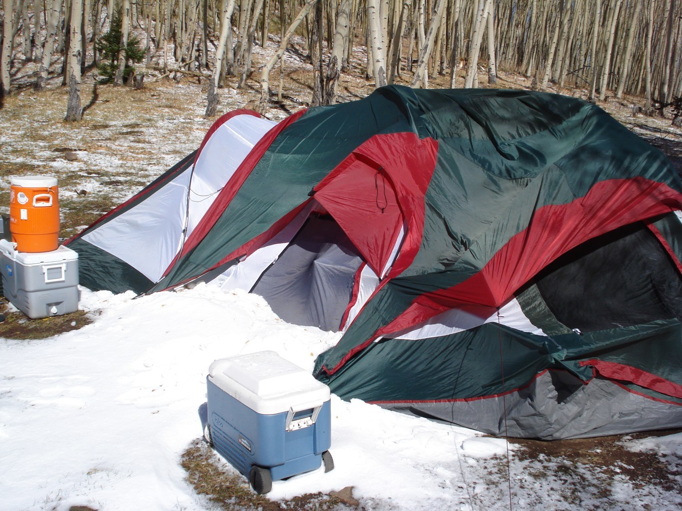 A large family dome tent collapsed to one side in a snowy aspen grove. The green and red fly is crumpled and sagging, with two coolers in front (photo brian gautreau, Wikimedia Commons, CC BY-SA 2.0)