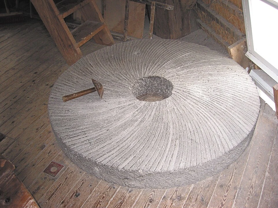 A round millstone lying on a wooden floor. It is a grey stone disc about an arm's length across with a round hole in the middle. Fine grooves curve outward from the centre to the edge across the whole face. A hammer with a steel bit lies on the stone