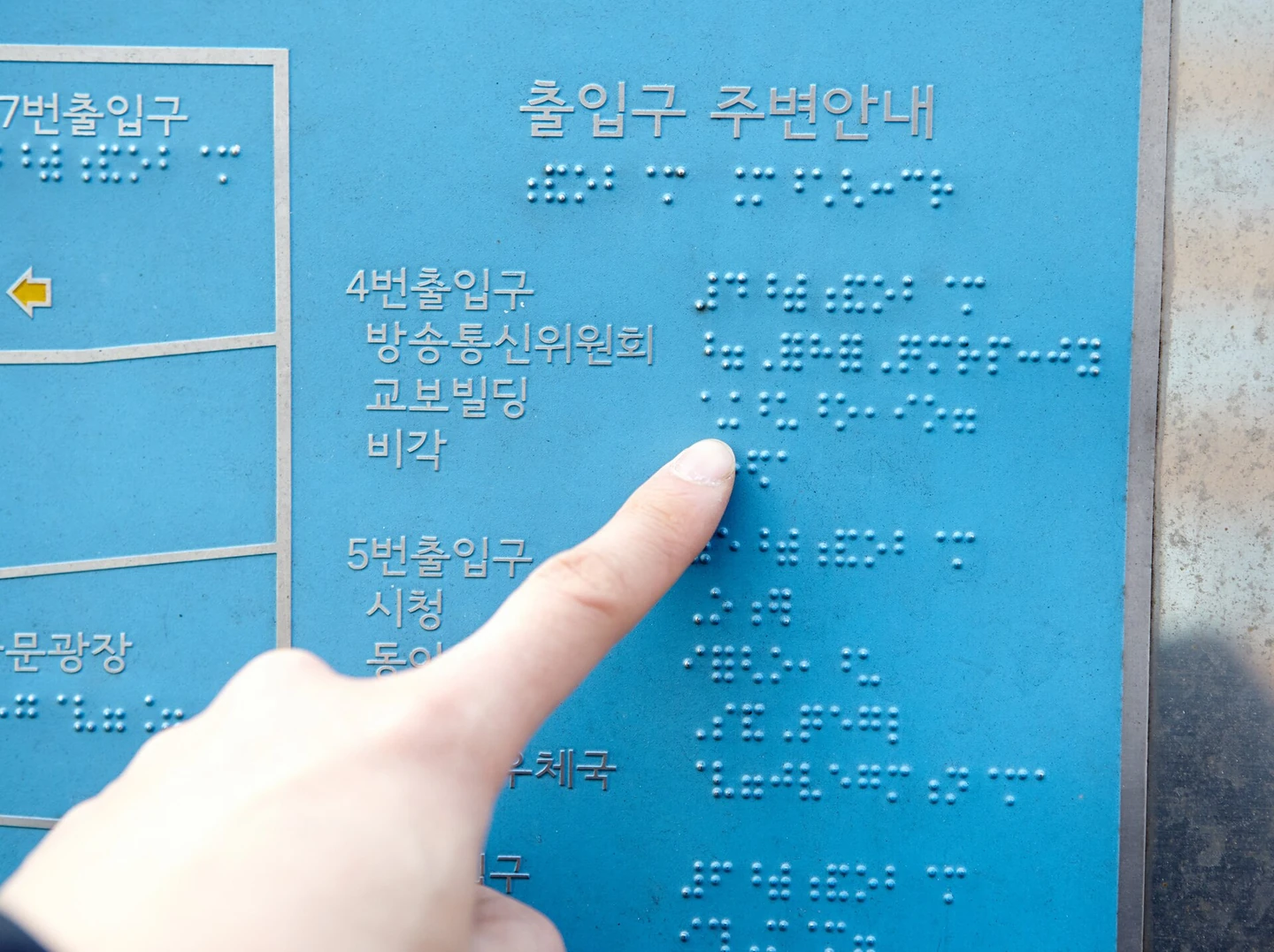 A sign near a subway station entrance. On a blue panel, Korean text such as "Exit 4, Korea Communications Commission" is printed, with raised braille dots just below it, and a finger is touching the braille (Photo: National Institute of Korean Language, Wikimedia Commons, CC BY-SA 2.0 KR)