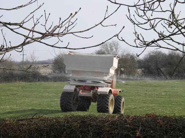 A tractor on green grass. From under the grey hopper at the back, white fertiliser granules are flung out in a wide fan to both sides
