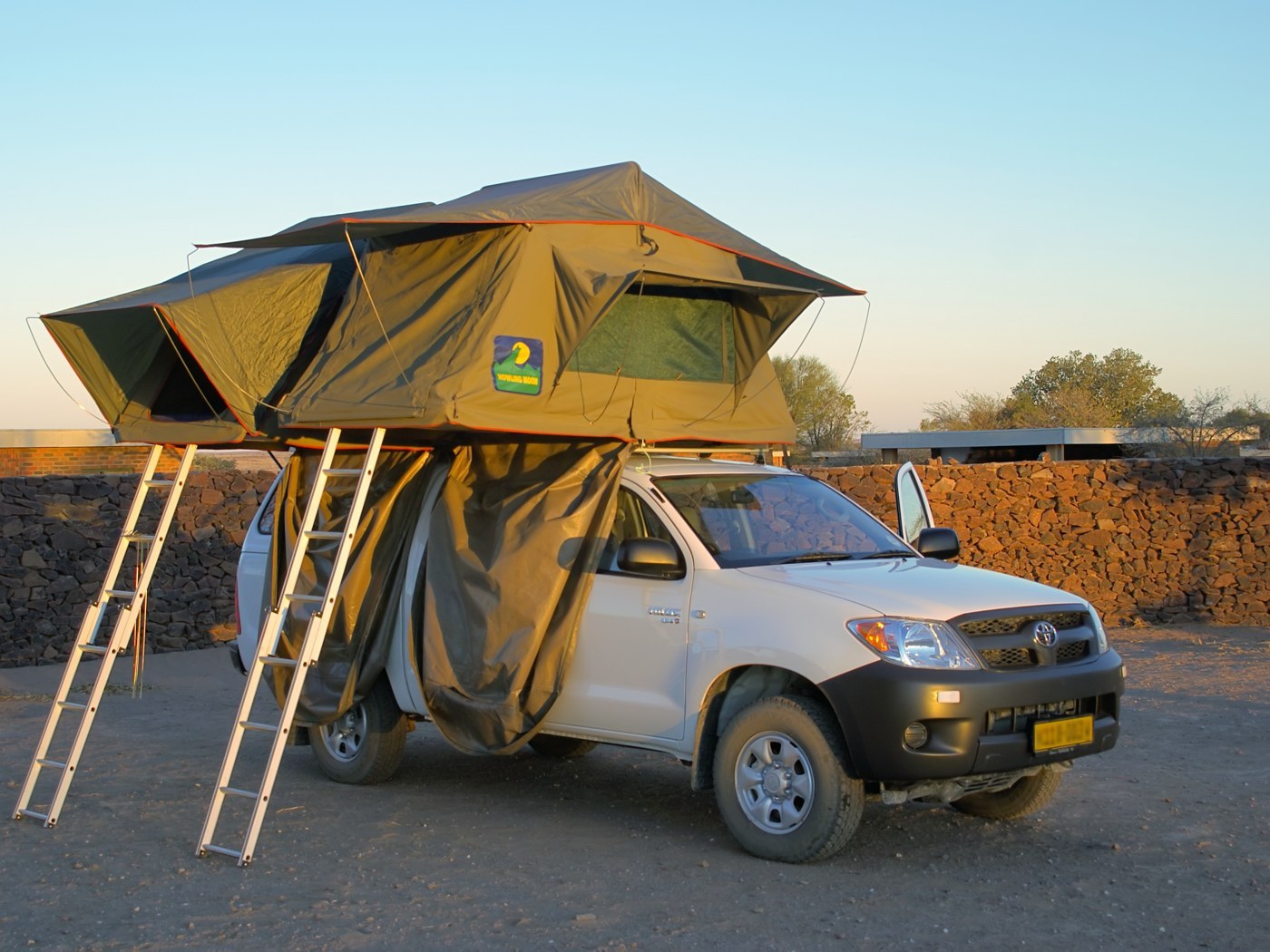 Two khaki fold-out rooftop tents opened on top of a white Toyota Hilux in a gravel yard in Namibia. Two aluminium ladders hold up the halves that fold out over the side of the car, with entry fabric hanging down below the tent (photo Hans Hillewaert, Wikimedia Commons, CC BY-SA 3.0)