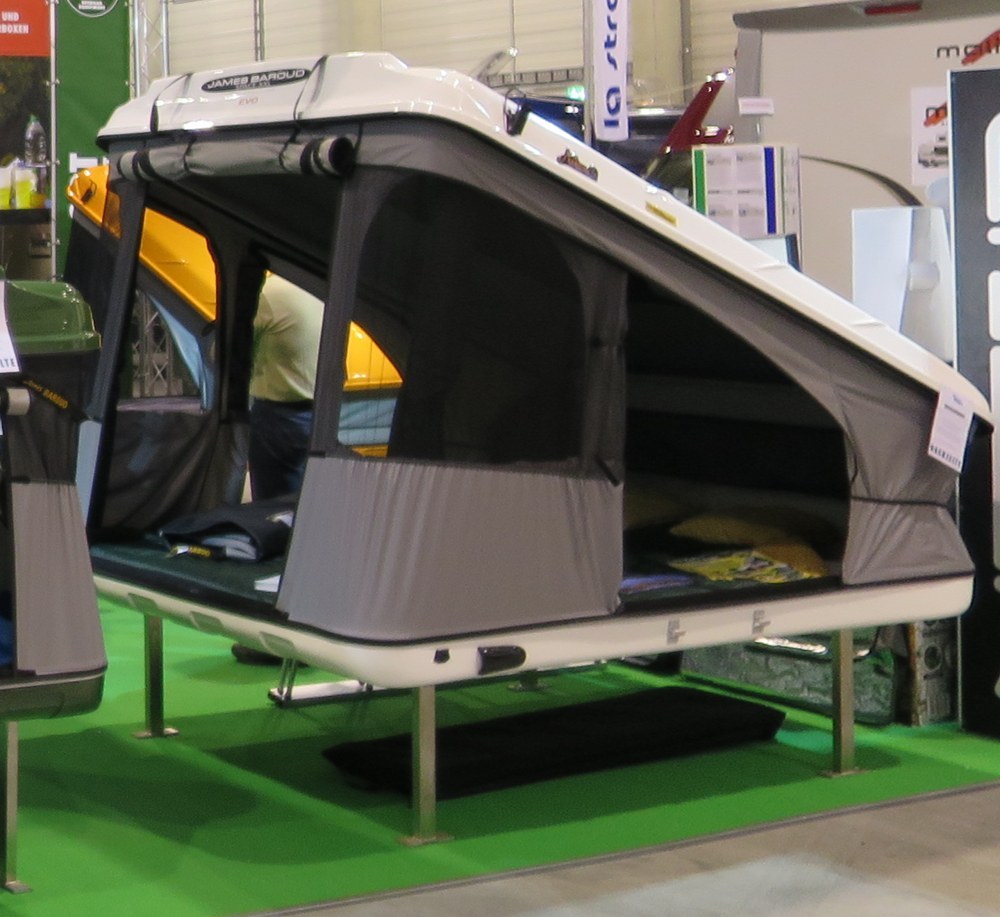 A white hard-shell wedge rooftop tent at a trade show. The lid rises from the hinge on the left toward the right, with grey fabric walls closing the gap between lid and base (photo Travelarz, Wikimedia Commons, CC BY-SA 3.0 pl, cropped)