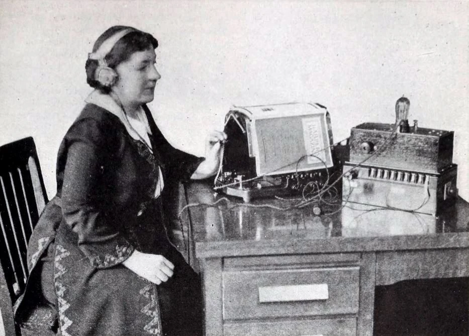 A 1922 magazine photograph. A woman wearing headphones sits at a desk, holding the handle of an optophone with a magazine propped on it. To the right is an amplifier box with a vacuum tube