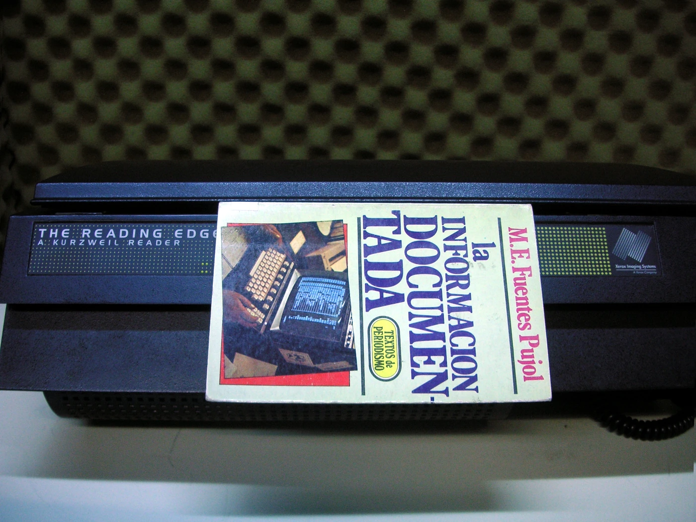 A book in Spanish stands on a black flatbed scanner. The front of the machine reads "THE READING EDGE, A KURZWEIL READER", with a "Xerox Imaging Systems" logo on the right